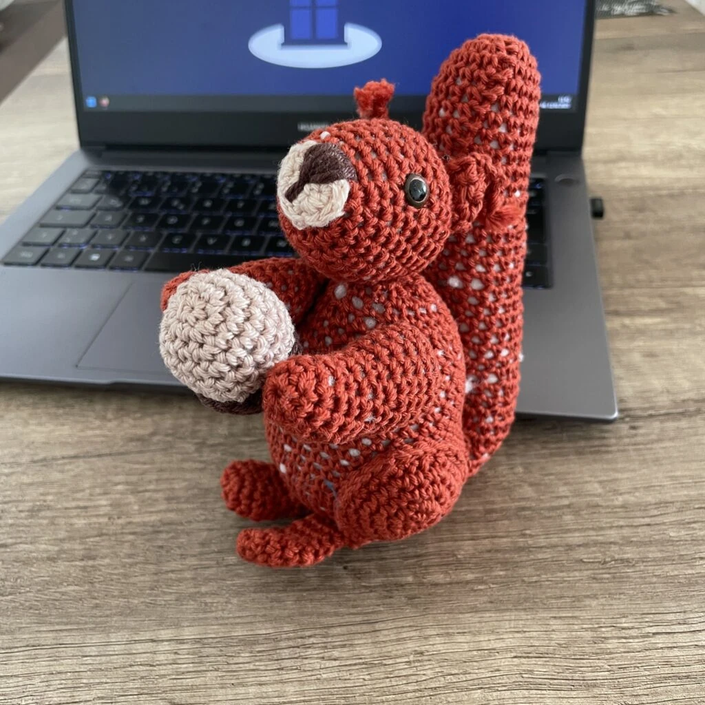 Gehäkeltes Eichhörnchen mit Nuss vor einem Laptop auf Holztisch