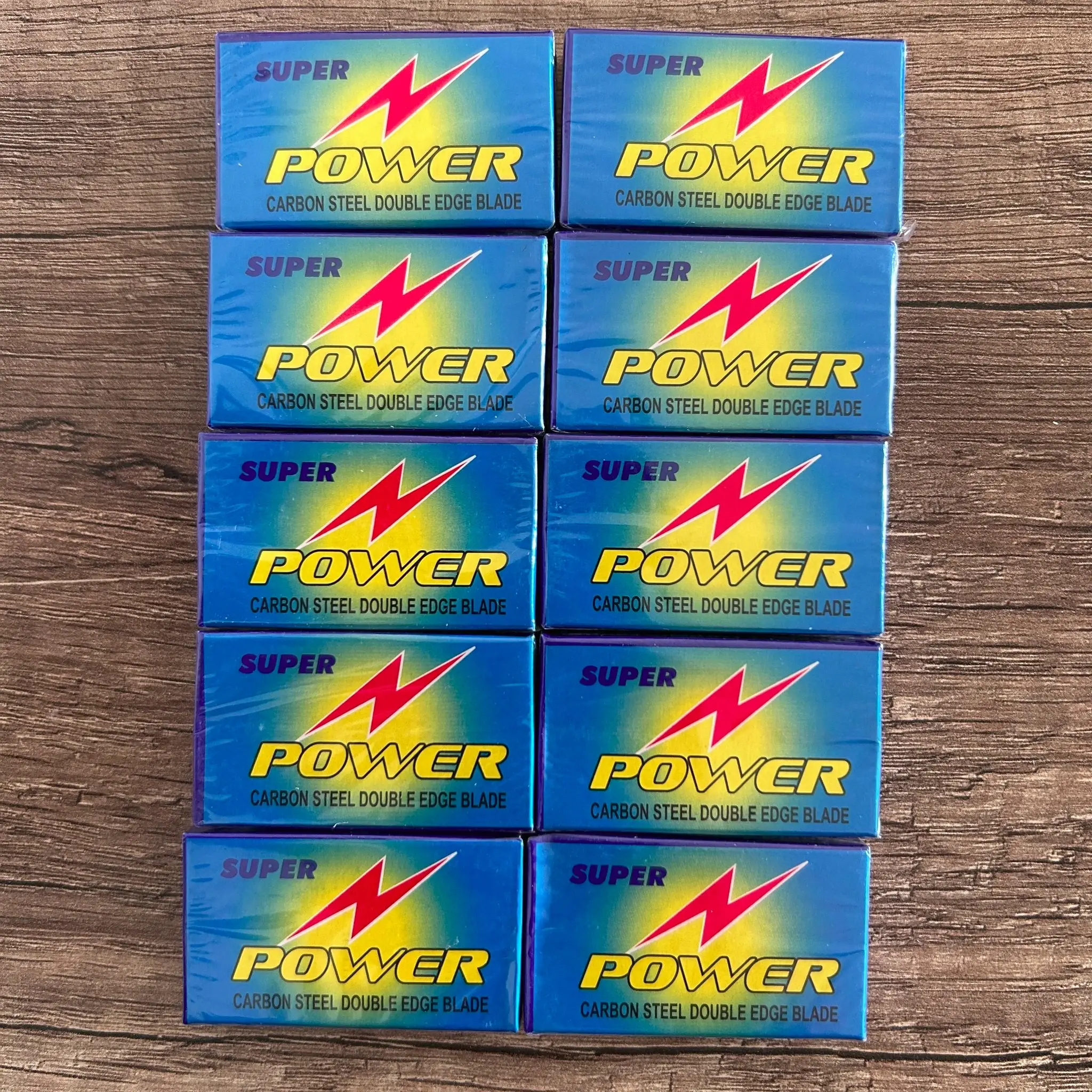 Zehn blaue Verpackungen mit der Aufschrift 'SUPER POWER CARBON STEEL DOUBLE EDGE BLADE' und rotem Blitzsymbol auf Holztisch angeordnet