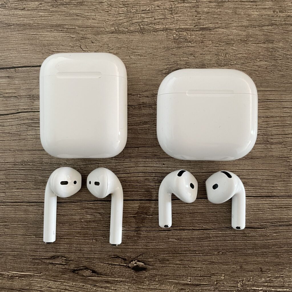 Das Bild zeigt zwei weiße AirPods-Ladehüllen und vier einzelne AirPods auf einer braunen Holztischplatte. Die Hüllen und die Ohrhörer liegen nebeneinander und zeigen deutlich die linke und rechte Seite. Links, die AirPods 2, rechts die AirPods 4.