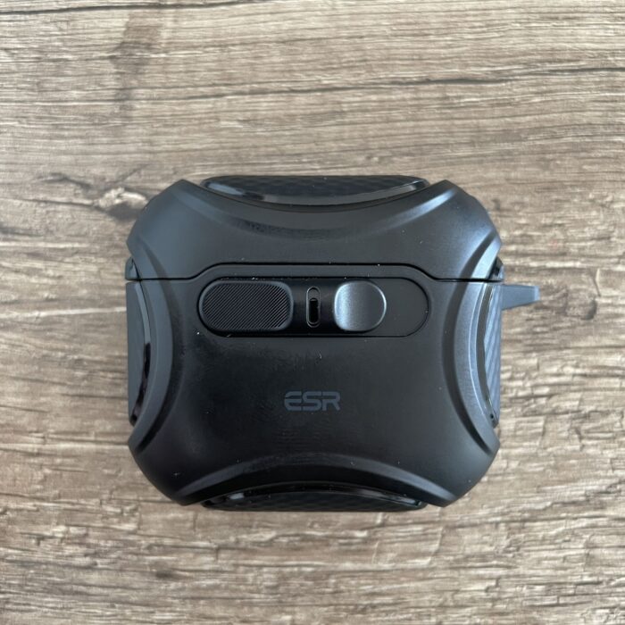 Das Bild zeigt eine schwarze Schutzhülle für AirPods, die auf einer Holztischplatte liegt. Die Hülle hat eine strukturierte Oberfläche und zwei Knöpfe auf der Oberseite. Der Herstellername „ESR“ ist in der Mitte der Hülle eingeprägt.
