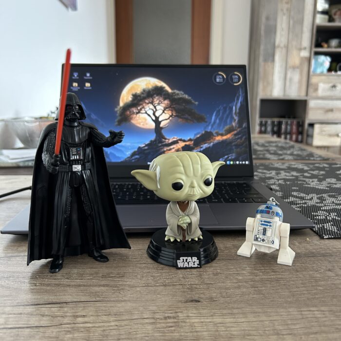 Das Bild zeigt drei Star Wars Figuren: Darth Vader mit rotem Lichtschwert, Yoda und R2-D2. Sie stehen auf einem Holztisch vor einem Laptop, auf dessen Bildschirm ein Bild eines Baumes vor einem Vollmond zu sehen ist.