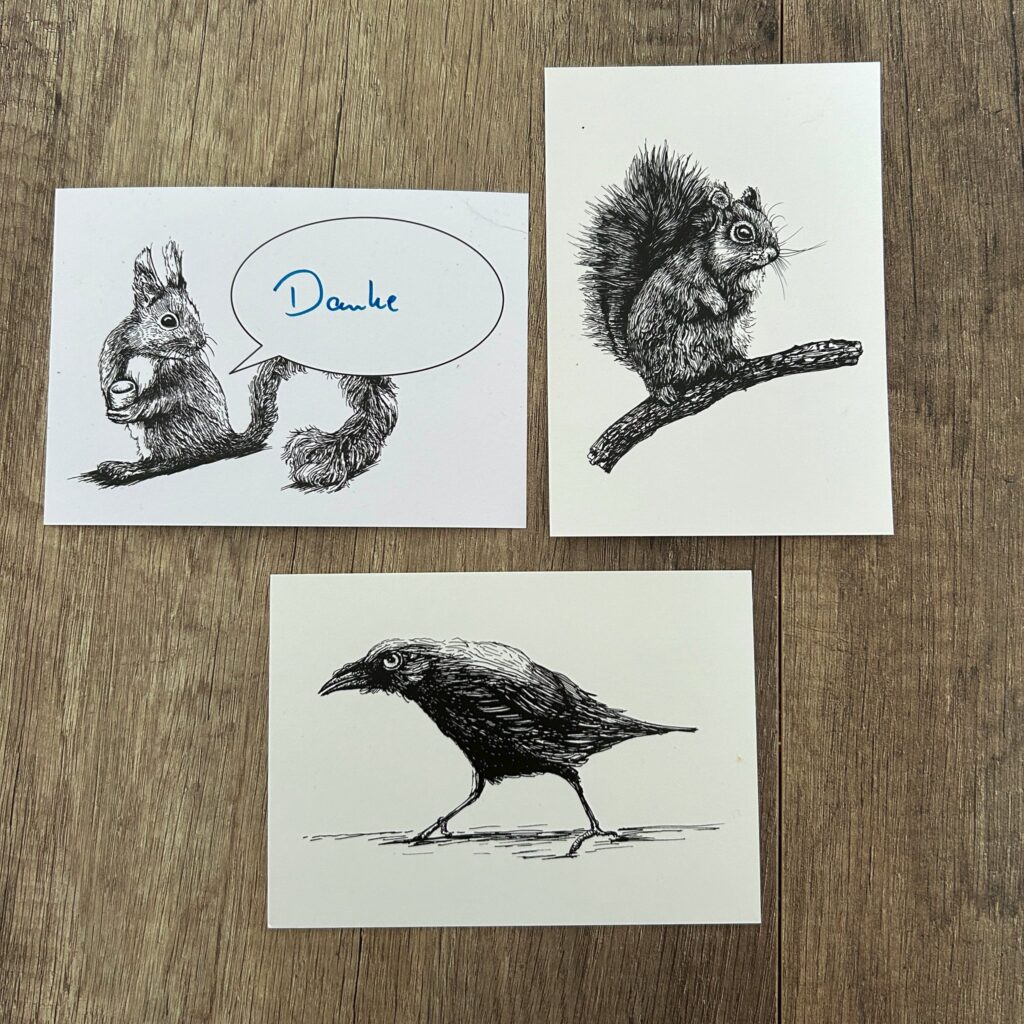 Das Bild zeigt drei Postkarten mit je einer Tuschezeichnung eines Tieres. Eine Karte zeigt einen Eichhörnchen, das eine Nuss hält und eine Sprechblase mit dem Wort „Danke“ enthält. Eine zweite Karte zeigt ein Eichhörnchen, das auf einem Ast sitzt. Die dritte Karte zeigt eine Krähe. Alle Zeichnungen sind in Schwarz-Weiß gehalten und wirken detailliert und realistisch. Die Postkarten liegen auf einem Holztisch.