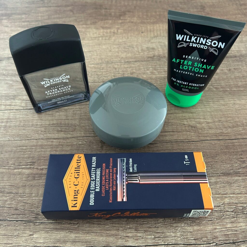 Das Bild zeigt eine Auswahl an Rasurprodukten auf einer Holzoberfläche. Zu sehen sind ein Flakon Wilkinson Sword After Shave Fragrance, eine Tube Wilkinson Sword Sensitive After Shave Lotion, eine Dose Rasierseife (ebenfalls von Wilkinson Sword) und einen King C. Gillette Double Edge Safety Razor Rasierhobel.