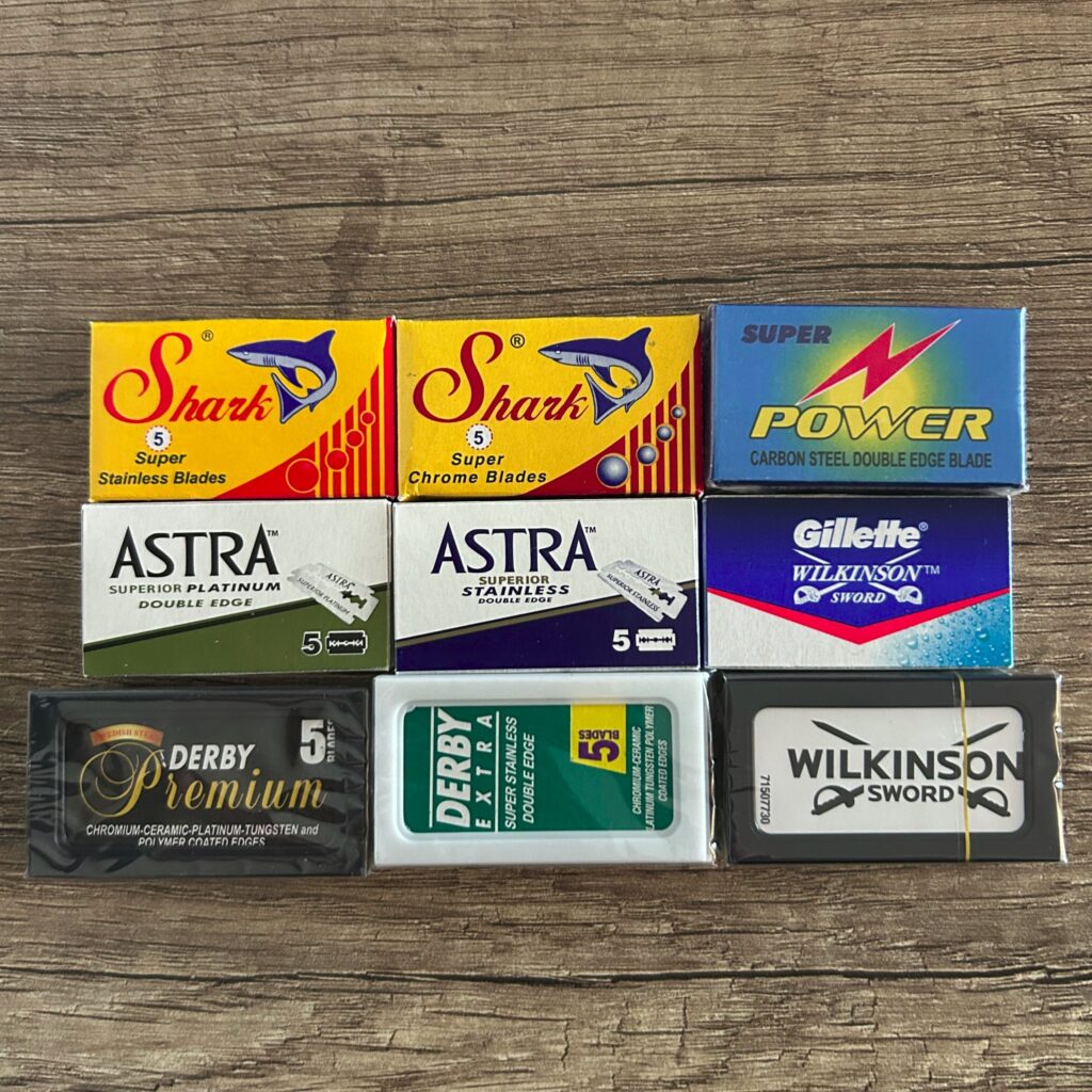 Das Bild zeigt neun Päckchen Rasierklingen verschiedener Marken. Die Marken sind Shark, Power, Astra, Gillette Wilkinson, Wilkinson Sword und Derby. Die Packungen sind auf einem Holztisch oder einer Holzoberfläche angeordnet.