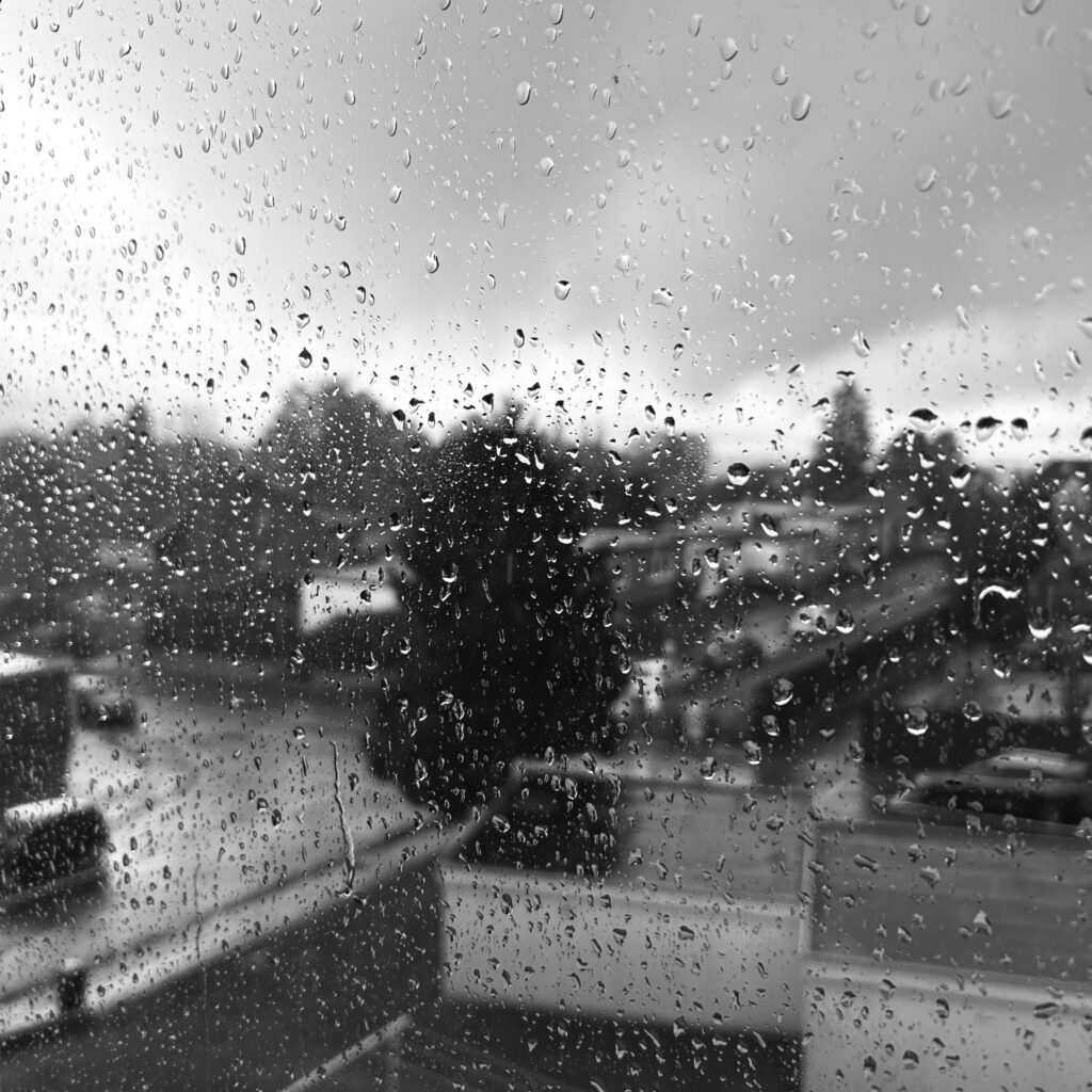 Das Bild zeigt eine schwarz-weiße Aufnahme durch eine regennasse Fensterscheibe. Im Vordergrund sind zahlreiche Wassertropfen auf dem Glas zu sehen, die das Licht brechen und die dahinterliegende Szene verzerrt darstellen. Im Hintergrund kann man verschwommen eine städtische oder vorstädtische Landschaft erkennen, möglicherweise mit Bäumen und Gebäuden. Die Szene wirkt etwas düster und melancholisch aufgrund des Regens und der Schwarz-Weiß-Farben.